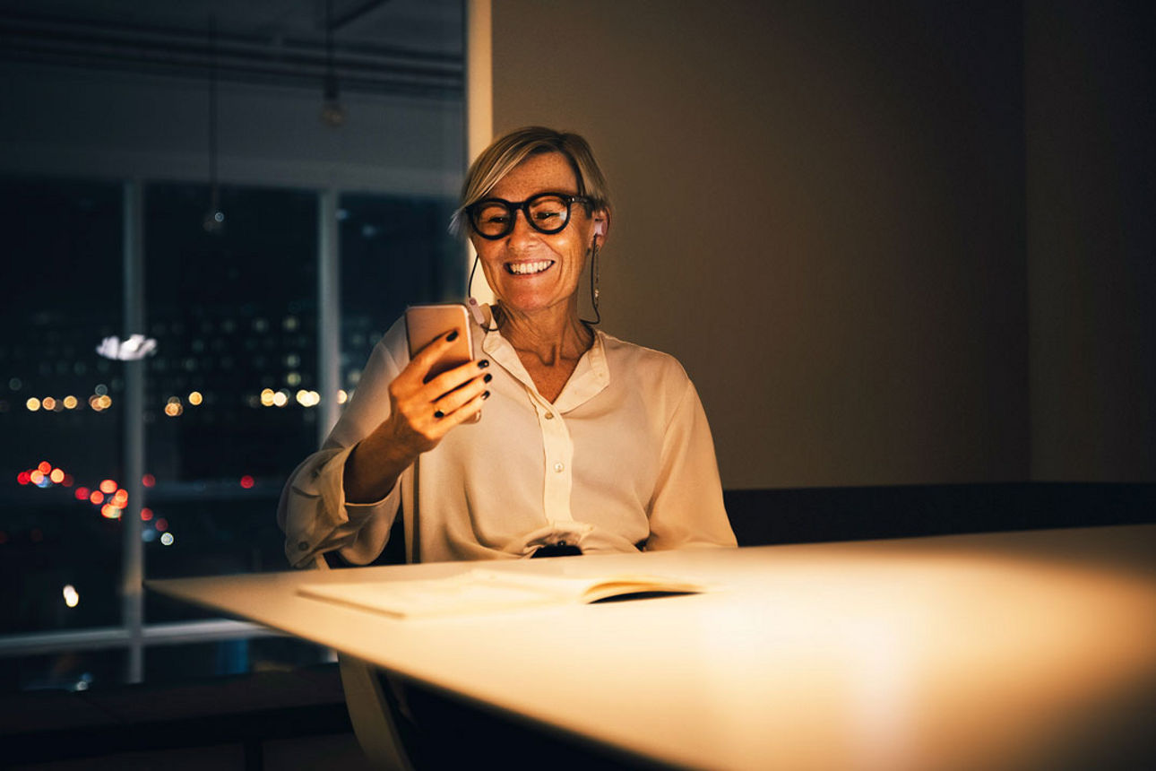 Frau sitzt an einem Tisch und schaut auf ihr Handy, sie lächelt dabei
