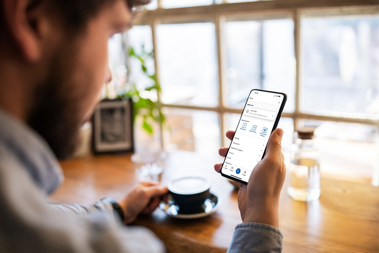 Junger Mann mit Kaffee in der einen und mit einem Smartphone in der anderen Hand, auf dem die BBBank-BankingApp geöffnet ist.