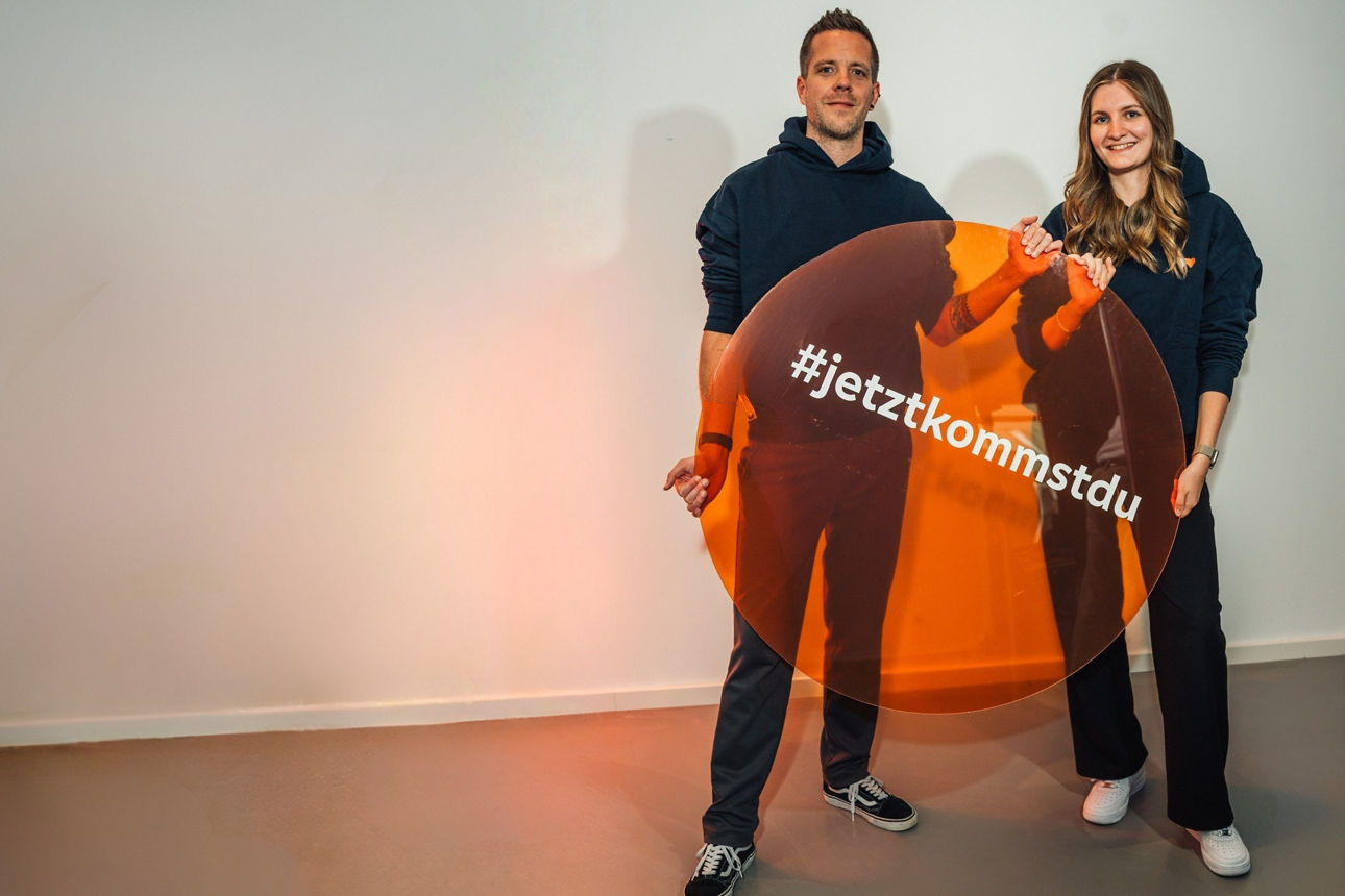  Zwei Personen in dunklen Hoodies halten ein rundes Schild mit der Aufschrift „#jetztkommstdu“.