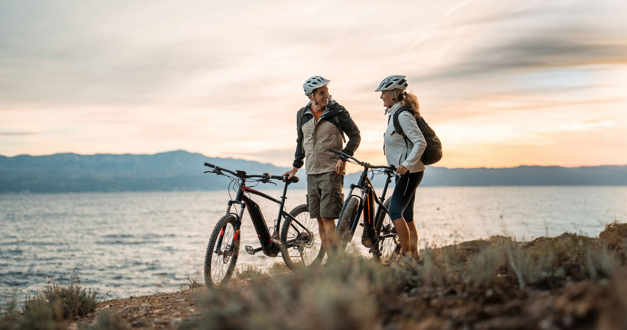 Ein Paar steht mit E-Bikes im sonnenuntergang an einer Klippe im Hintergrund sieht man das Meer.
