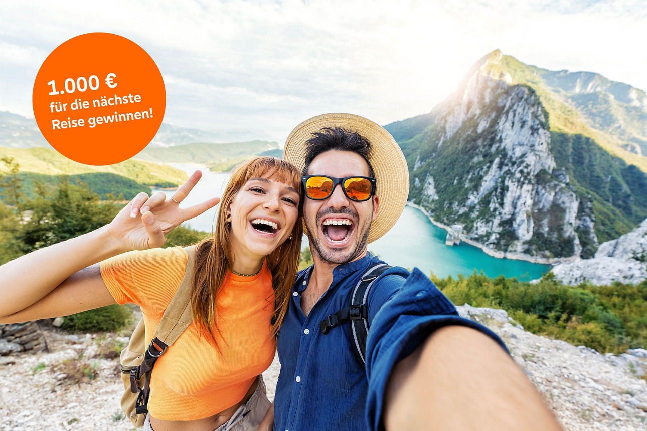 Ein lachendes Paar macht ein Selfie vor einer Bergkulisse mit Fluss. Oben links ein orangefarbener Kreis mit dem Text ‚1.000 € für die nächste Reise gewinnen!‘.