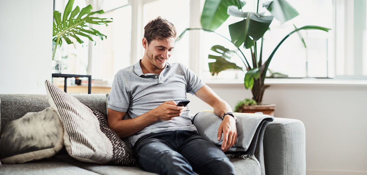 Ein Mann auf einem grauen Sofa mit einem Handy in der Hand. Im Hintergrund sind Pflanzen.
