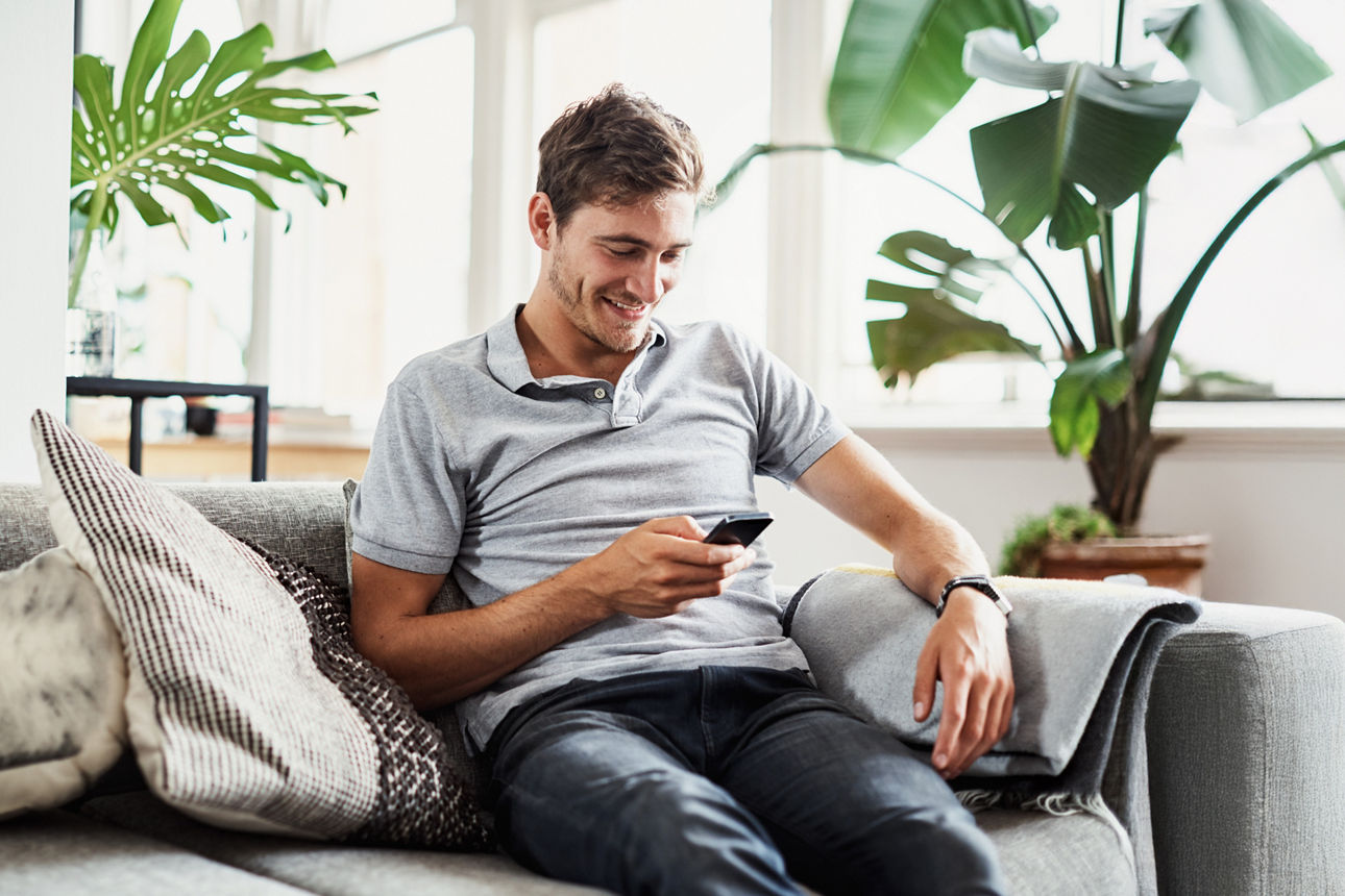 Ein Mann auf einem grauen Sofa mit einem Handy in der Hand. Im Hintergrund sind Pflanzen.