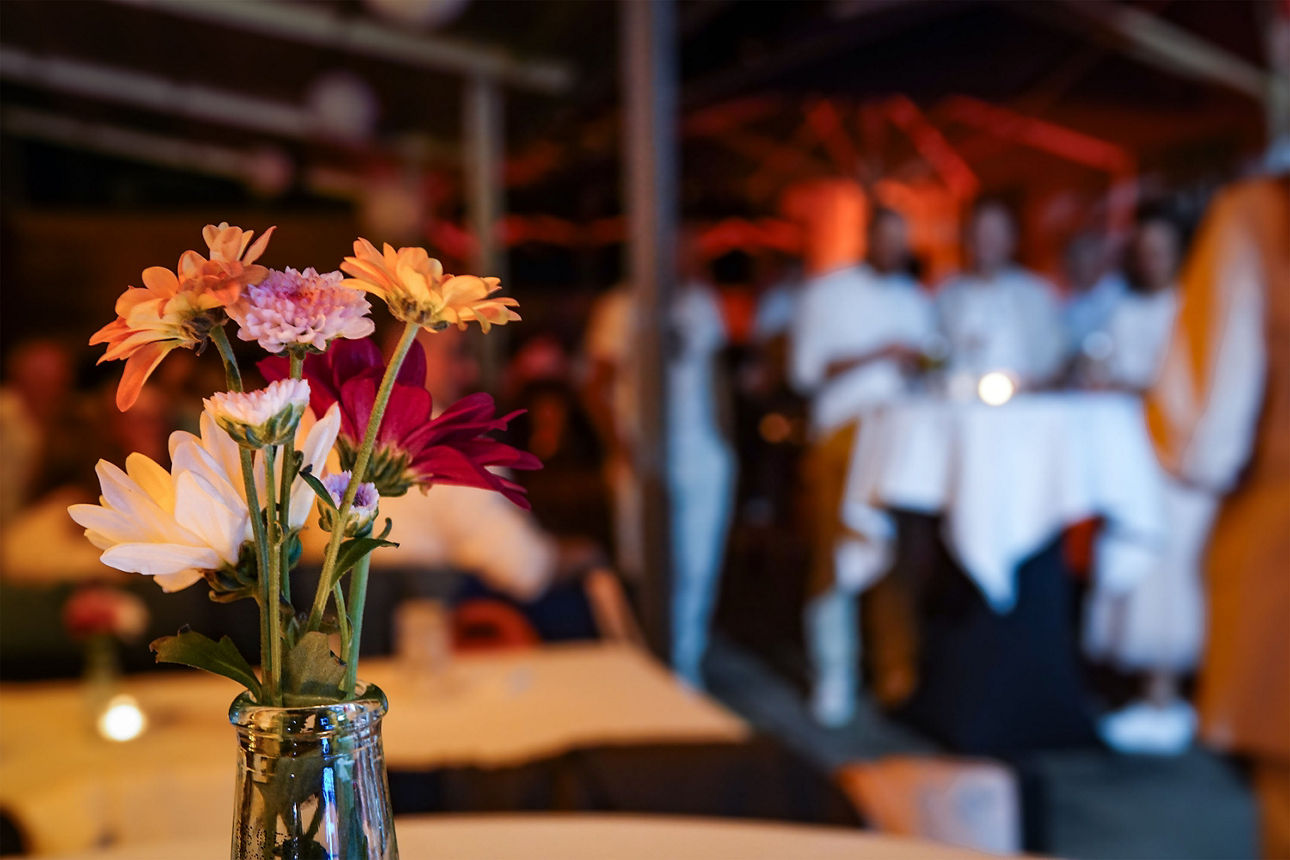 Eine Blumenvase auf dem Tisch, im Hintergrund stehen Menschen bei einer Veranstaltung.