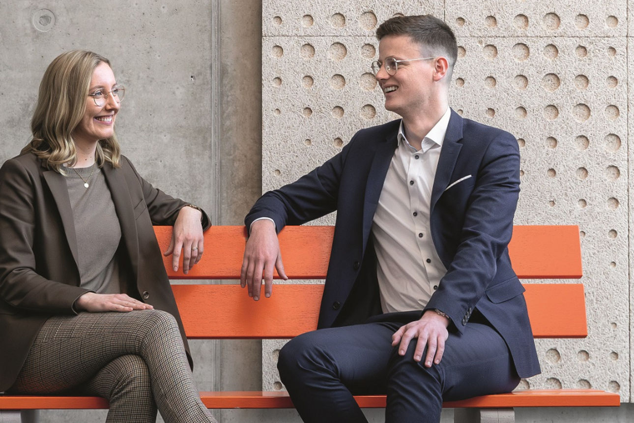 Ein junger Mann und eine junge Frau sitzen lächelnd auf einer Parkbank