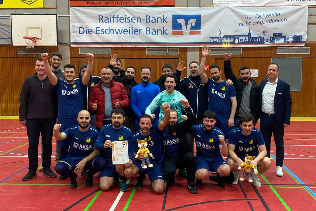 Handballmannschaft mit Pokalen und Betreuern in Turnhalle vor Raiffeisen-Banner