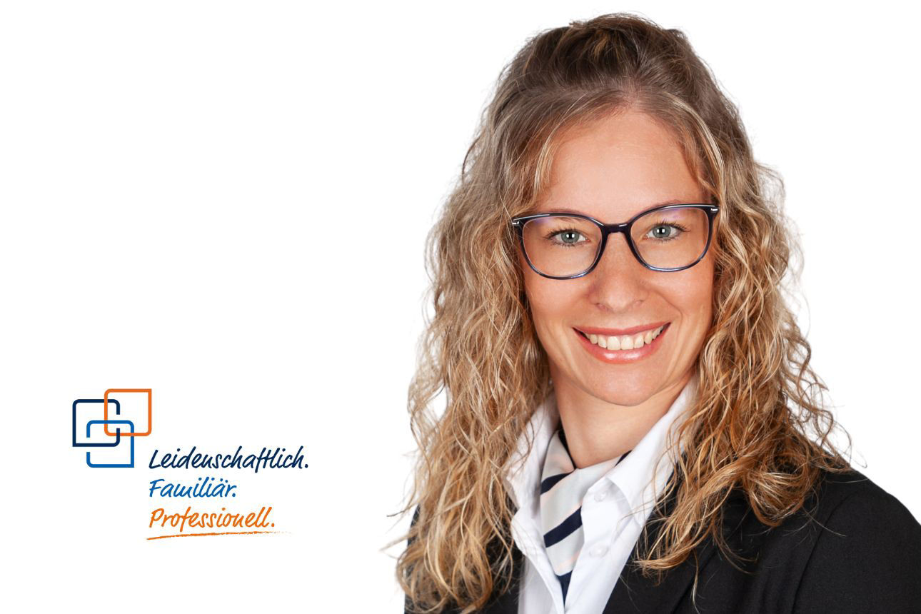 Heidi Zacher, eine Frau mit langen, lockigen blonden Haaren und Brille, lächelt freundlich und trägt ein dunkles Sakko über einem gestreiften weiß-blauen Oberteil.
