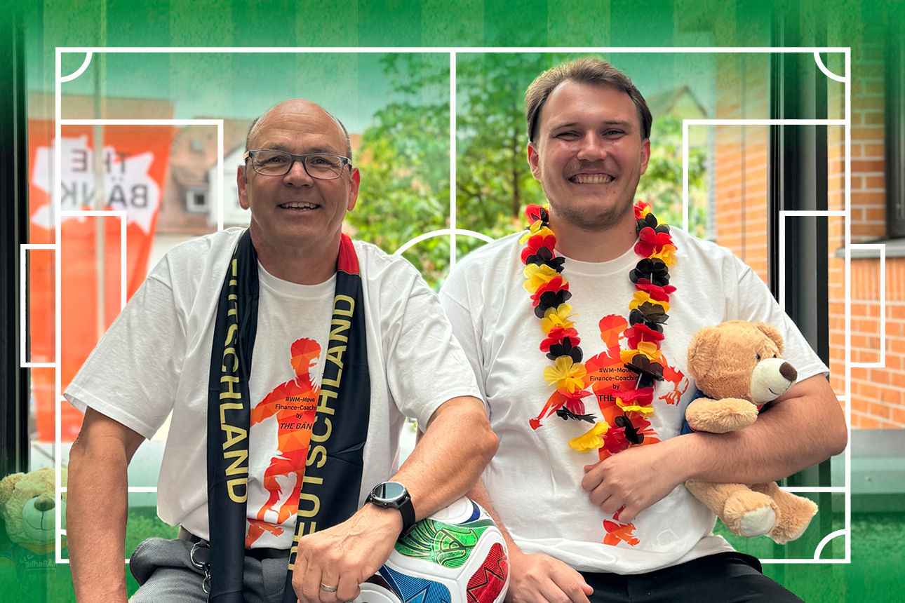 Sie sehen zwei Mitarbeiter mit WM-Utensilien, die nebeneinander sitzen.