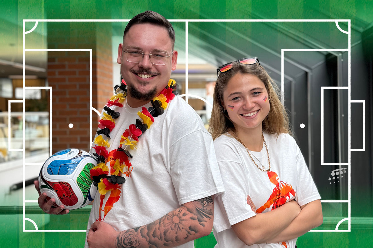 Sie sehen 2 Mitarbeiter, die mit WM-Utensilien nebeneinander stehen.