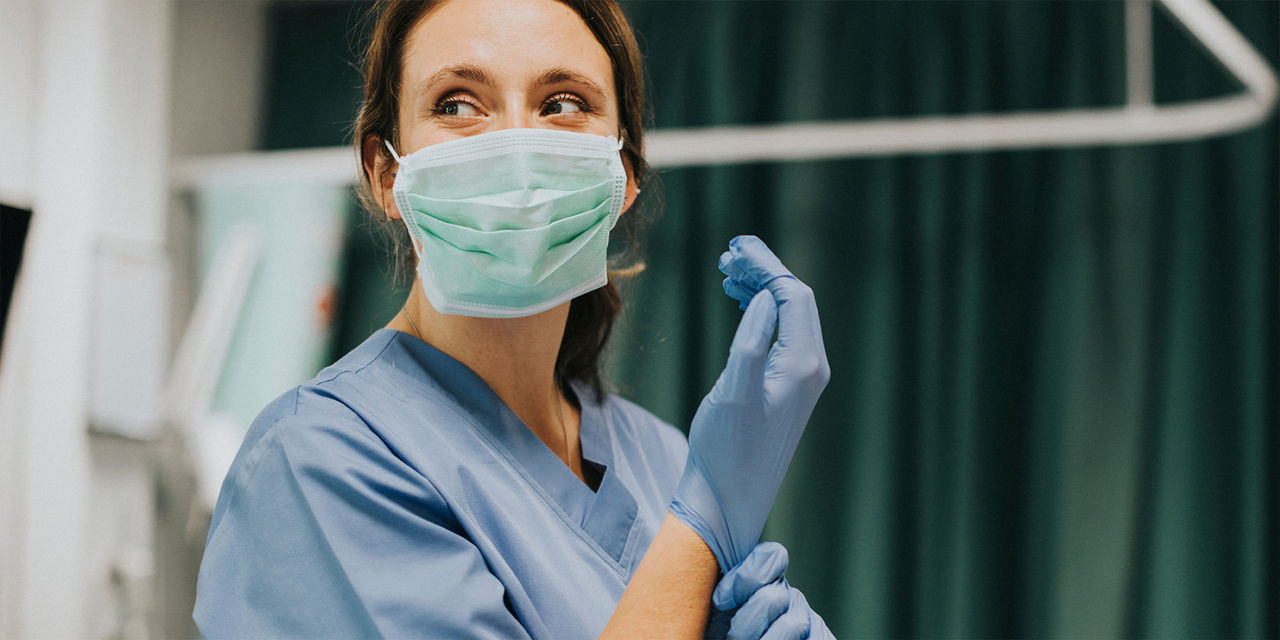 Eine medizinische Fachkraft in blauer Schutzkleidung, mit Gesichtsmaske und Handschuhen, die sich gerade einen Handschuh anzieht, blickt zur Seite