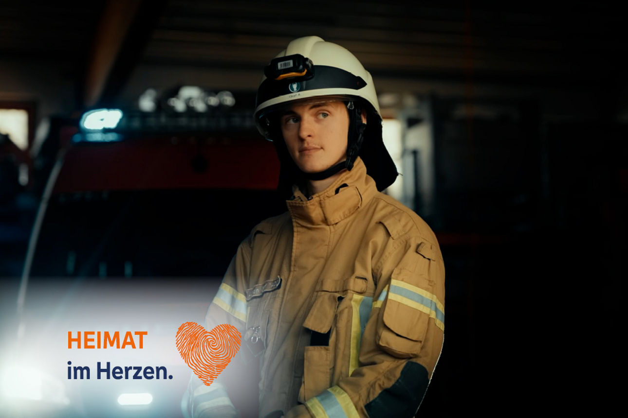 Feuerwehrmann in Uniform vor Feuerwehrauto, Text: HEIMAT im Herzen