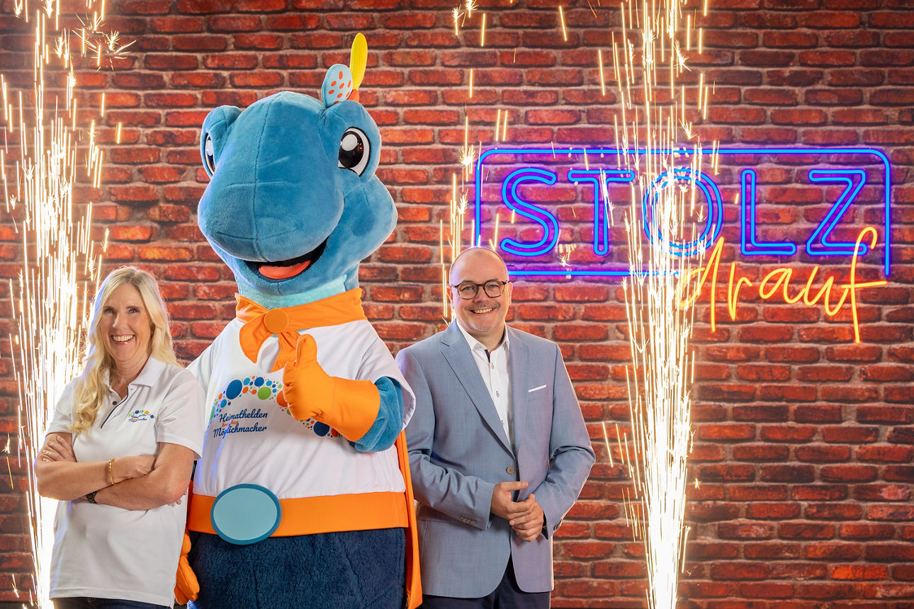 Mann und Frau mit einem großen Dinosaurier in der Mitte im Hintergrund zwei Feuerfontänen 