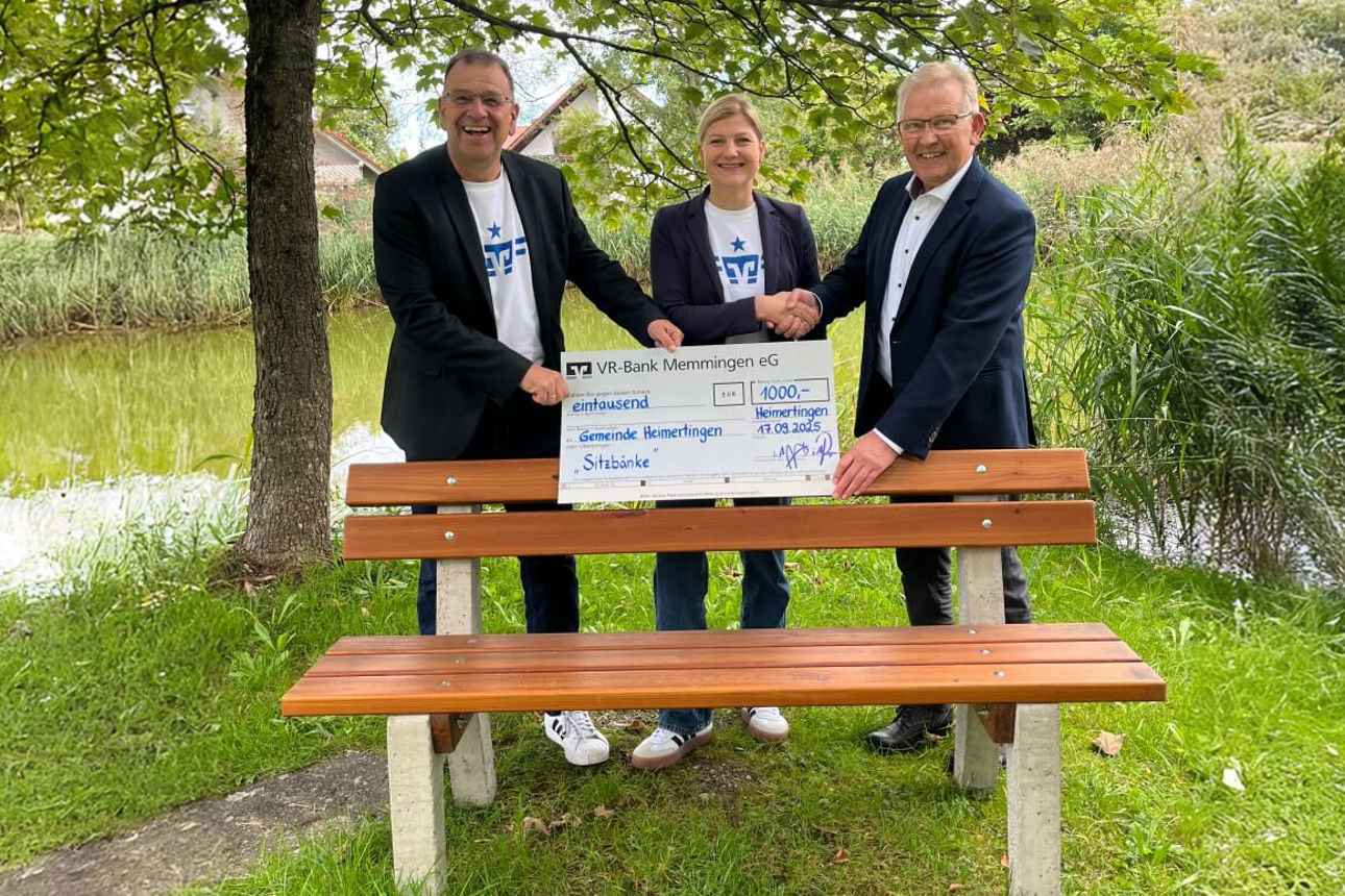 Bankmitarbeiter schütteln Bürgermeister die Hand und stehen mit Spendenscheck vor Sitzbank