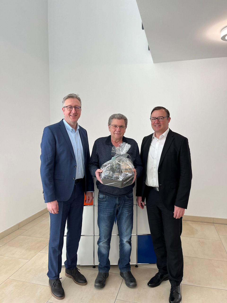 Das Bild zeigt drei Männer in einem hellen Gebäude, wobei einer von ihnen einen Geschenkkorb hält.