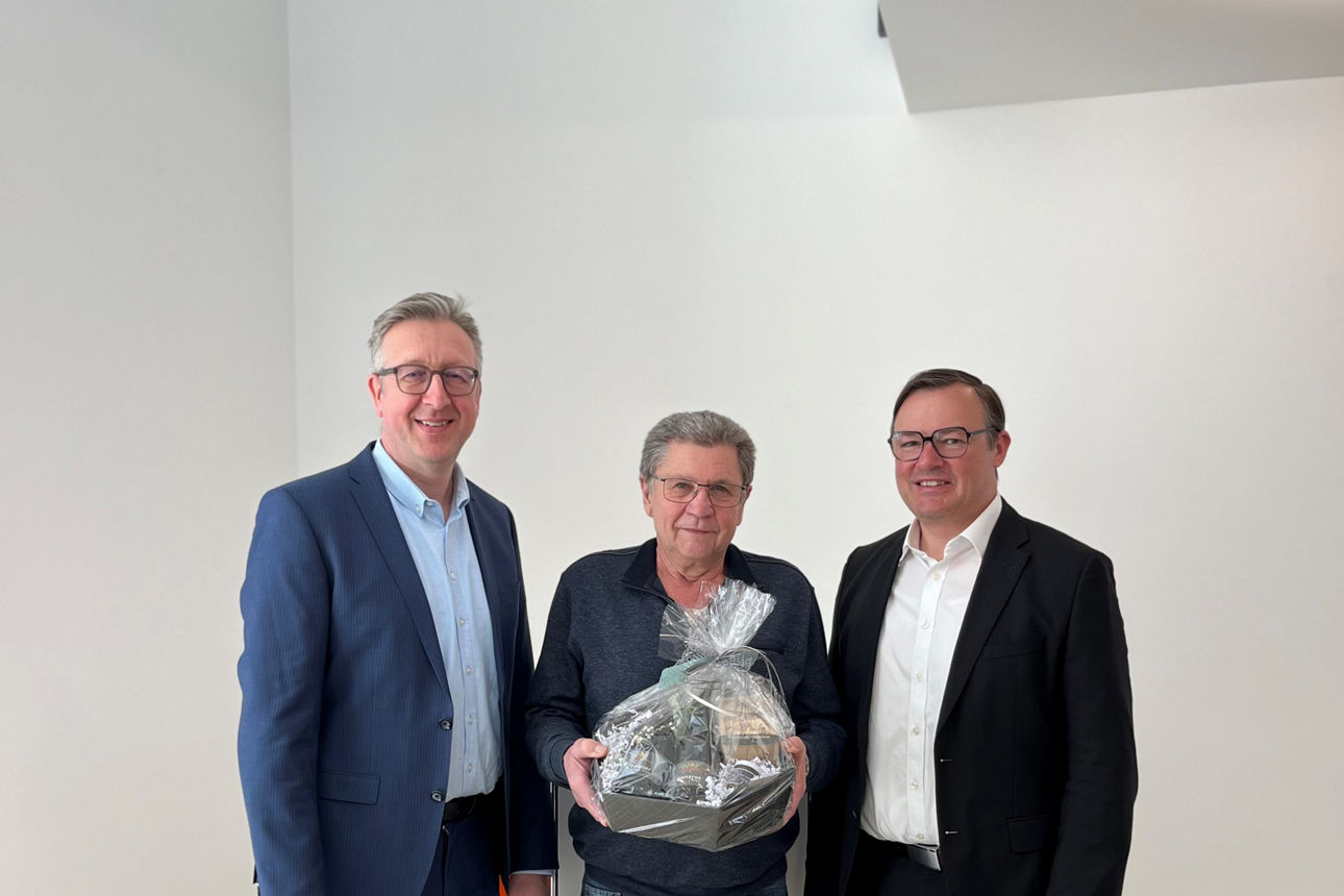 Das Bild zeigt drei Männer in einem hellen Gebäude, wobei einer von ihnen einen Geschenkkorb hält.