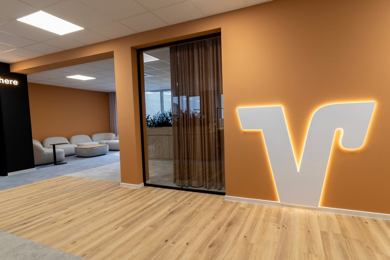 Innenraum mit hellbrauner Wand, beleuchtetem großen V-förmigem Logo, Holzboden und Sitzbereich mit Sesseln im Hintergrund