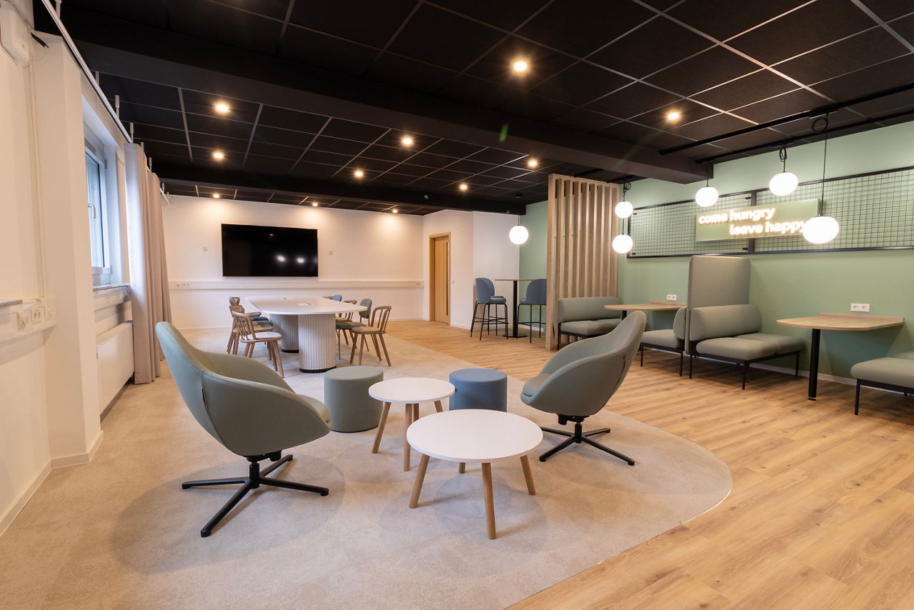 Moderner, offener Aufenthaltsraum mit runden Tischen, Sesseln auf Teppich und Sitzbank mit Hockern vor Wand mit Leuchtschrift