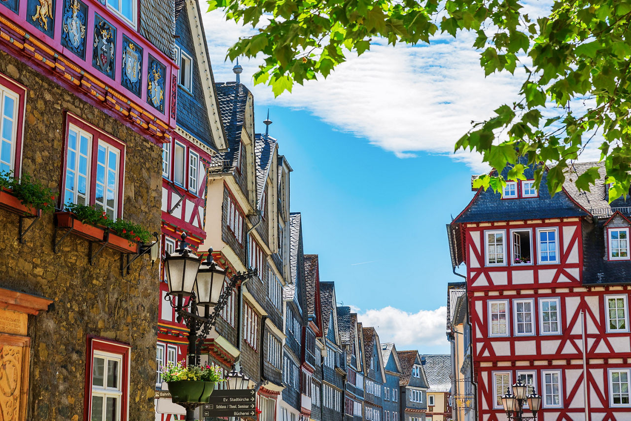 Blick in eine farbenfrohe Fachwerkhaus-Straße der Altstadt von Herborn, geschmückt mit Blumenkästen, unter blauem Himmel und grünen Baumkronen.