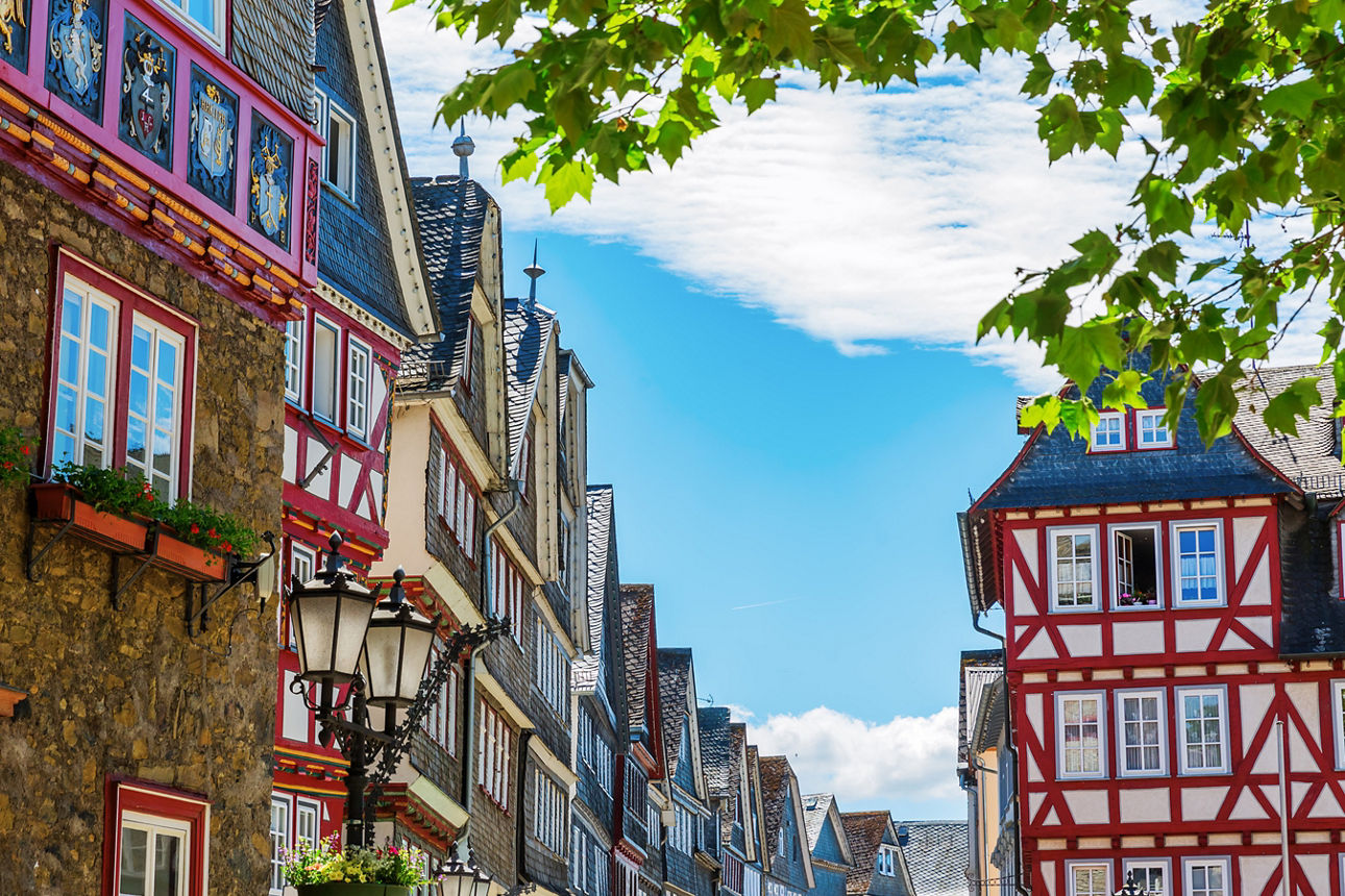 Blick in eine farbenfrohe Fachwerkhaus-Straße der Altstadt von Herborn, geschmückt mit Blumenkästen, unter blauem Himmel und grünen Baumkronen.