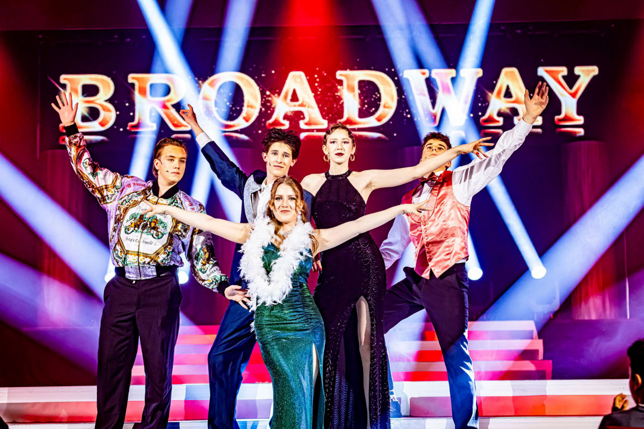 Bühne mit Schriftzug Broadway, 5 glamourös gekleidete Menschen mit gehobenen Armen am Ende eines Liedes