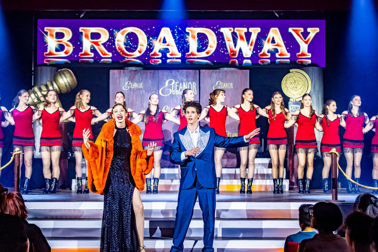 Zwei Darsteller singen vor einer Gruppe Tänzerinnen, alle gekleidet in Show-Kostümen