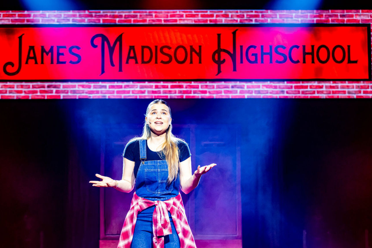 junge Frau singt vor einem Schild James Madison Highschool, leger gekleidet in T-Shirt und Jeans Latzhose