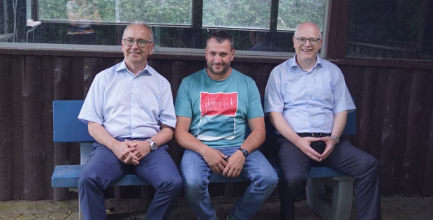 Drei Männer sitzen auf einer blauen Bank vor einem Holzgebäude.