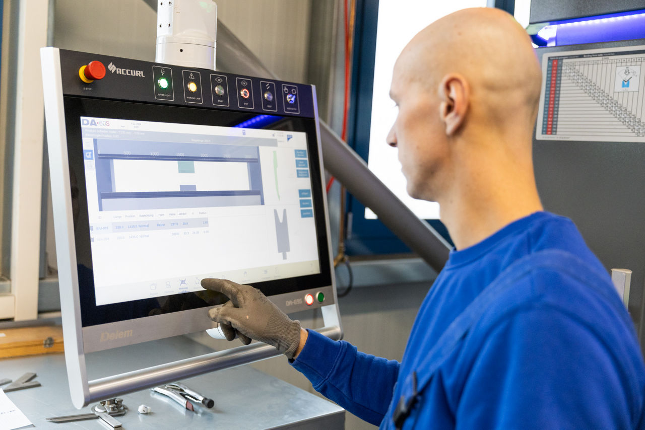 Interaktion mit der Benutzeroberfläche einer CNC‑Abkantpresse bei der „Herrlich GmbH“, bei der Biegeparameter für die Blechbearbeitung eingestellt werden. Sichtbar sind das Steuerungspanel und die grafische Darstellung des anstehenden Abkantvorgangs im Produktionsumfeld