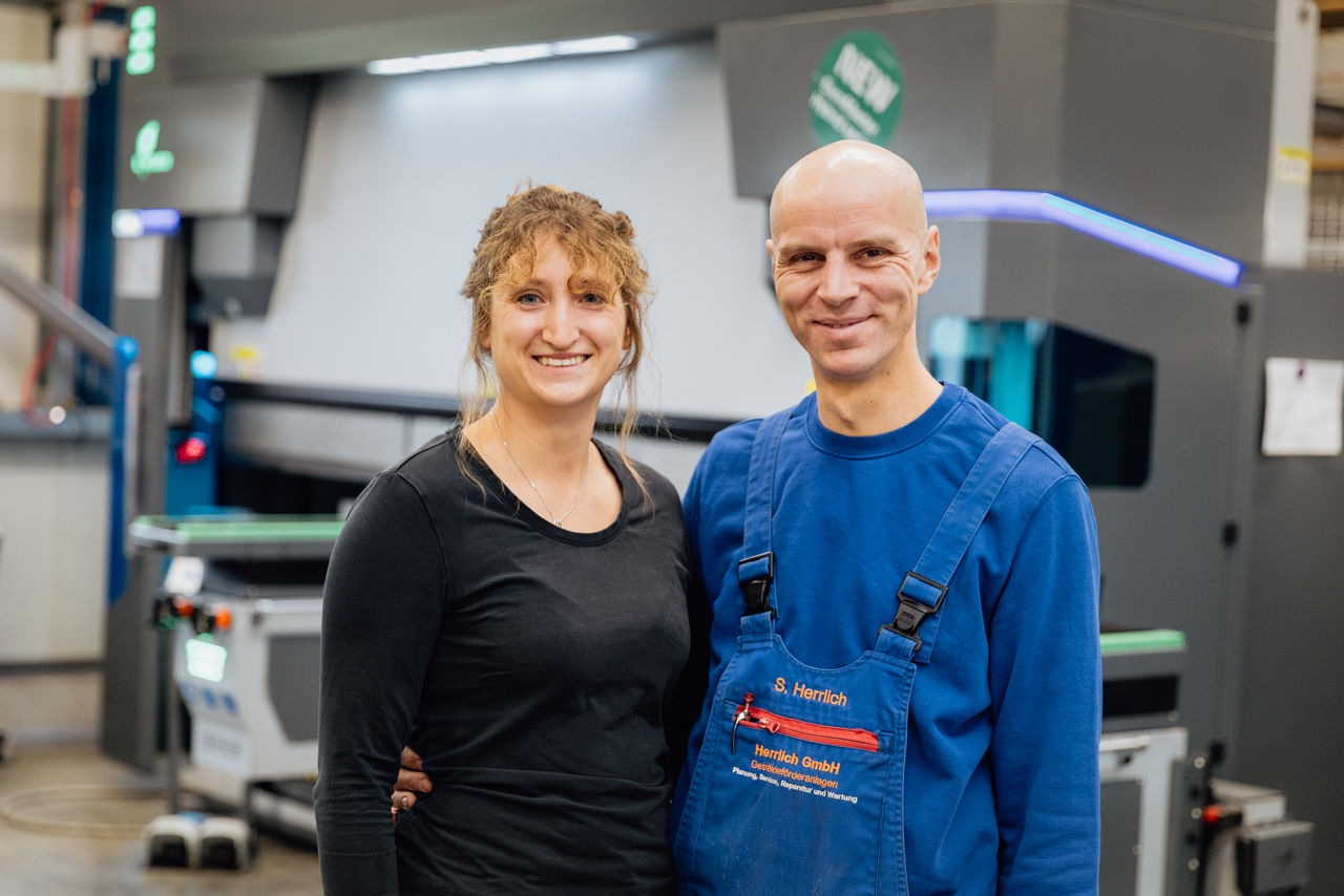 Zwei Personen stehen im Produktionsumfeld der „Herrlich GmbH“ vor einer industriellen Abkantpresse. Die Aufnahme zeigt die Metallbearbeitungsanlage im Fertigungsbereich mit Fokus auf maschinelle Blechbearbeitung und industrielle Produktionsprozesse