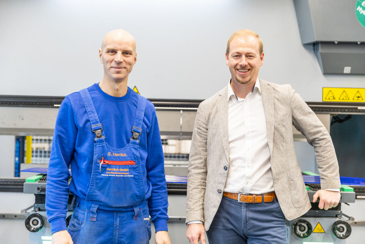 Sebastian Herrlich und Berater Florian Abschütz stehen im Produktionsbereich der „Herrlich GmbH“ vor einer Abkantpresse. Die Aufnahme zeigt den Metallbearbeitungsbetrieb mit Fokus auf industrielle Blechbearbeitung und maschinelle Fertigungsprozesse