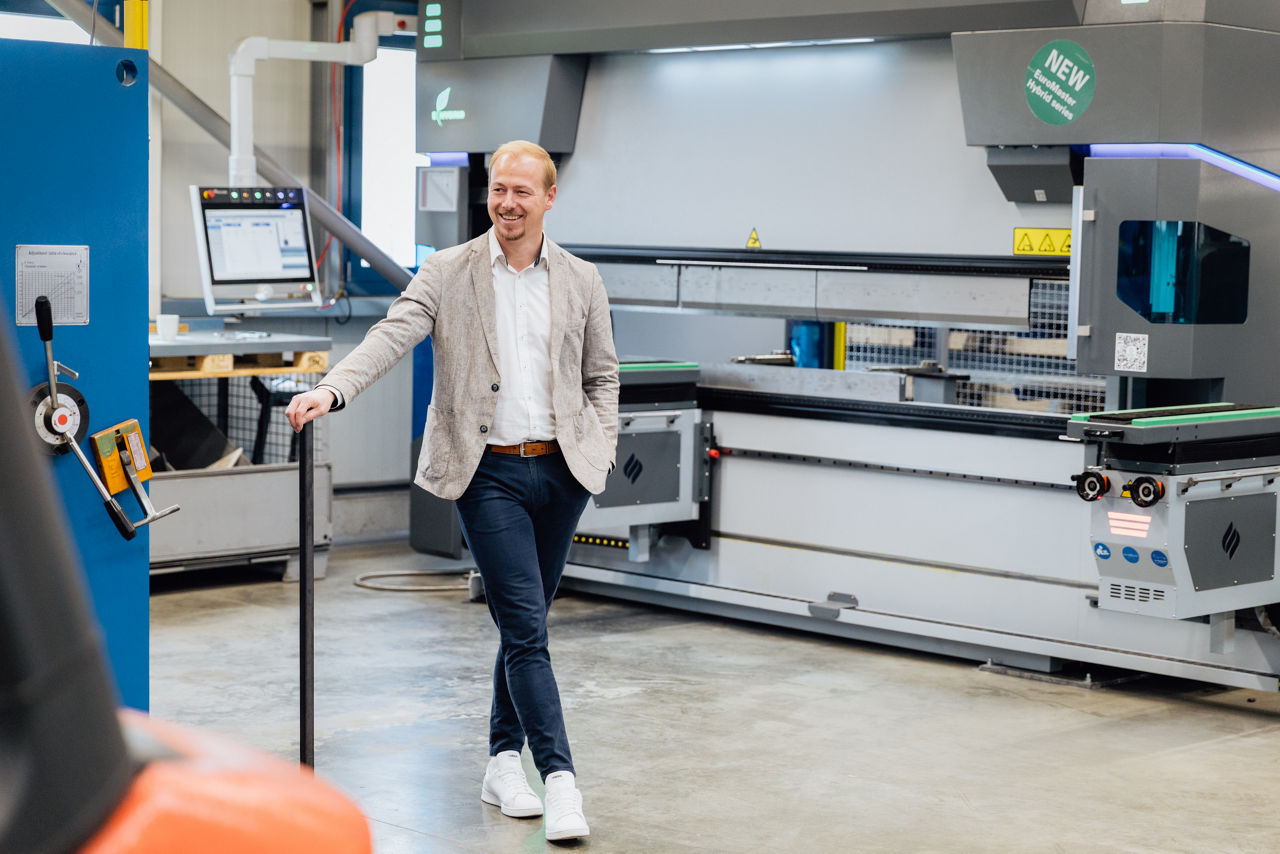 Berater Florian Abschütz befindet sich im Produktionsbereich der „Herrlich GmbH“ vor einer CNC‑Abkantpresse während eines laufenden Fertigungsablaufs. Die Aufnahme zeigt den industriellen Metallbearbeitungsprozess mit Fokus auf präzise Blechbearbeitung und moderne CNC‑Technologie im Produktionsumfeld