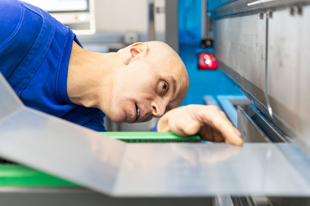 Präzise Kontrolle eines Blechteils an einer CNC‑Abkantpresse im Produktionsbereich der „Herrlich GmbH“ während des Fertigungsprozesses. Die Aufnahme zeigt den laufenden Abkantvorgang und die Qualitätsüberprüfung im Rahmen der industriellen Blechbearbeitung