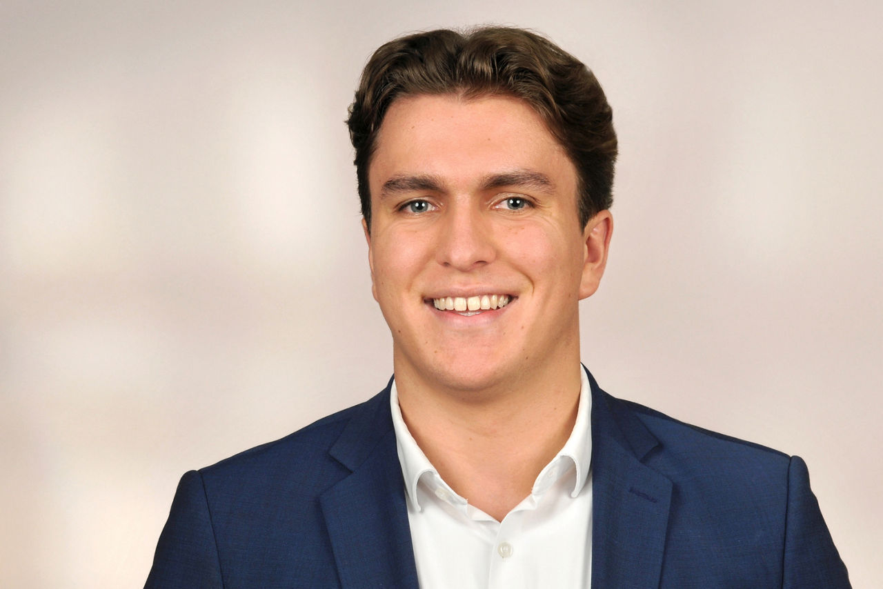 Mann mit braunen Haaren trägt weißen Hemd und dunkelblauen Anzug vor neutralem Hintergrund