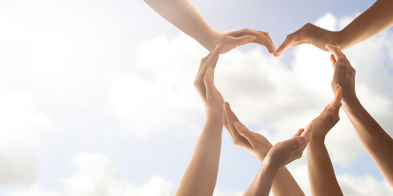 Mehrere Hände formen ein Herzsymbol vor hellem Himmel und stehen für Einheit und Zusammenarbeit.