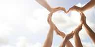Mehrere Hände formen ein Herzsymbol vor hellem Himmel und stehen für Einheit und Zusammenarbeit.