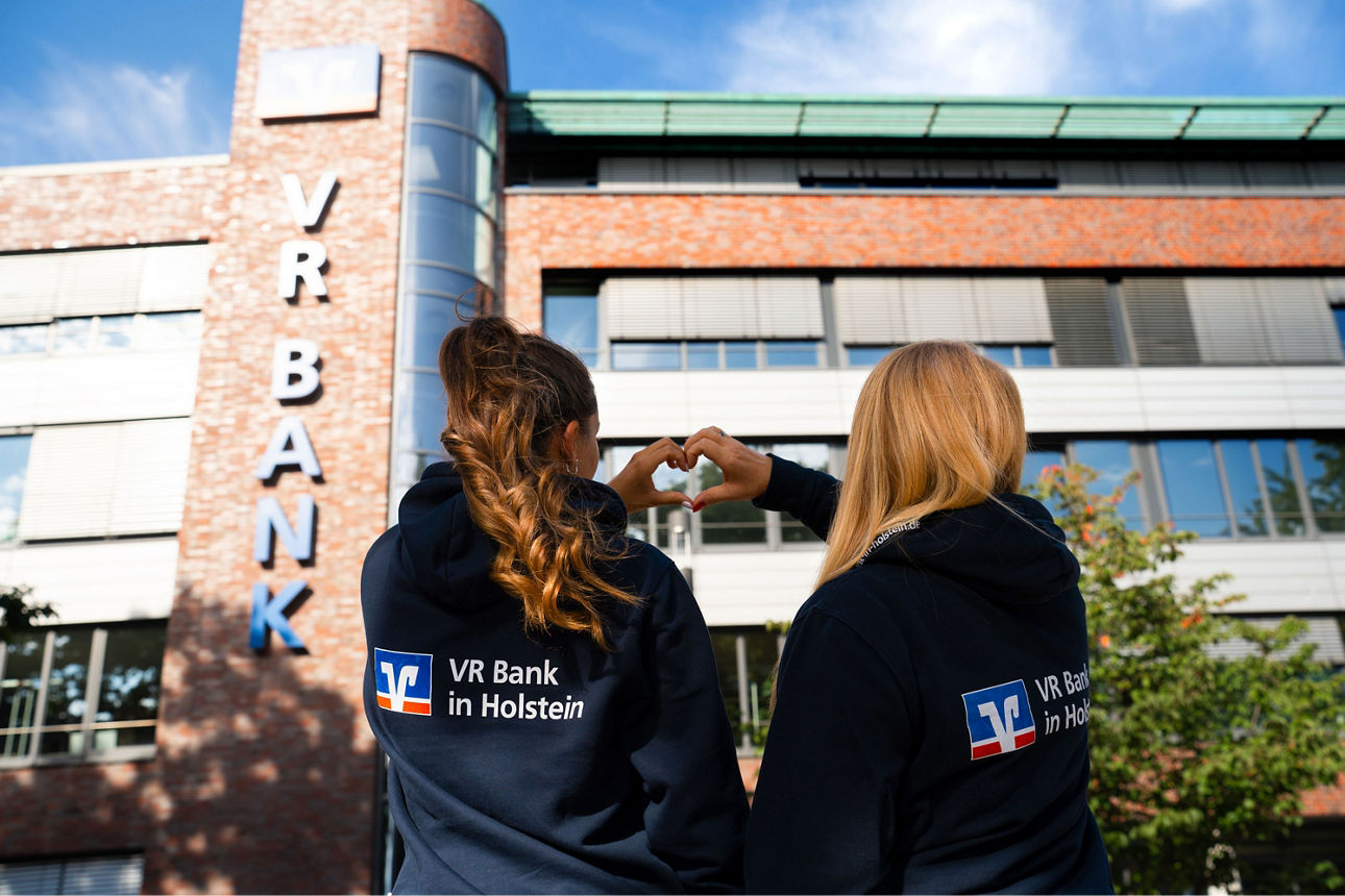 2 Mitarbeiterinnnen schauen aufs Bankgebäude und formen ein Herz mit den Händen