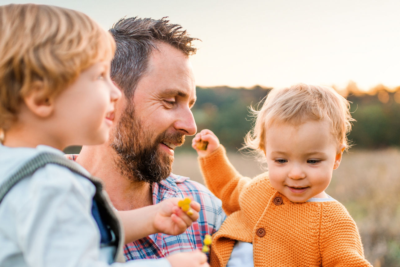 Ein Vater steht mit seinen 2 Söhnen auf dem Arm vor einer Wiese