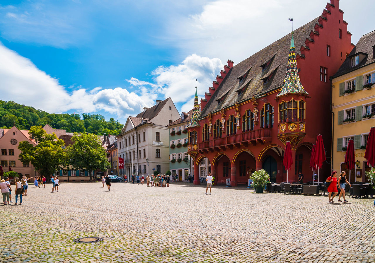 Gepflasterter Platz mit Menschen, historischen Häusern und einem auffälligen roten Gebäude mit Erker.