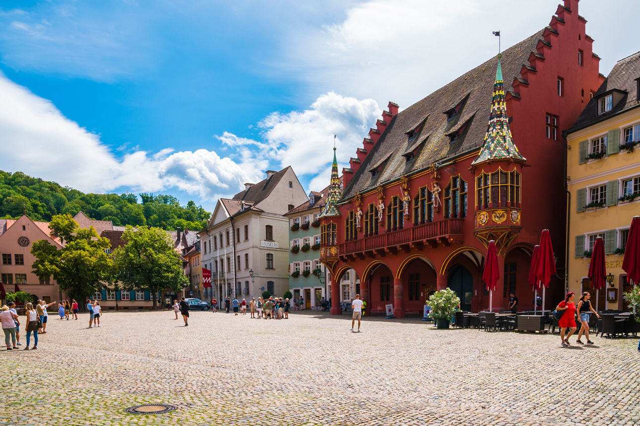 Gepflasterter Platz mit Menschen, historischen Häusern und einem auffälligen roten Gebäude mit Erker.