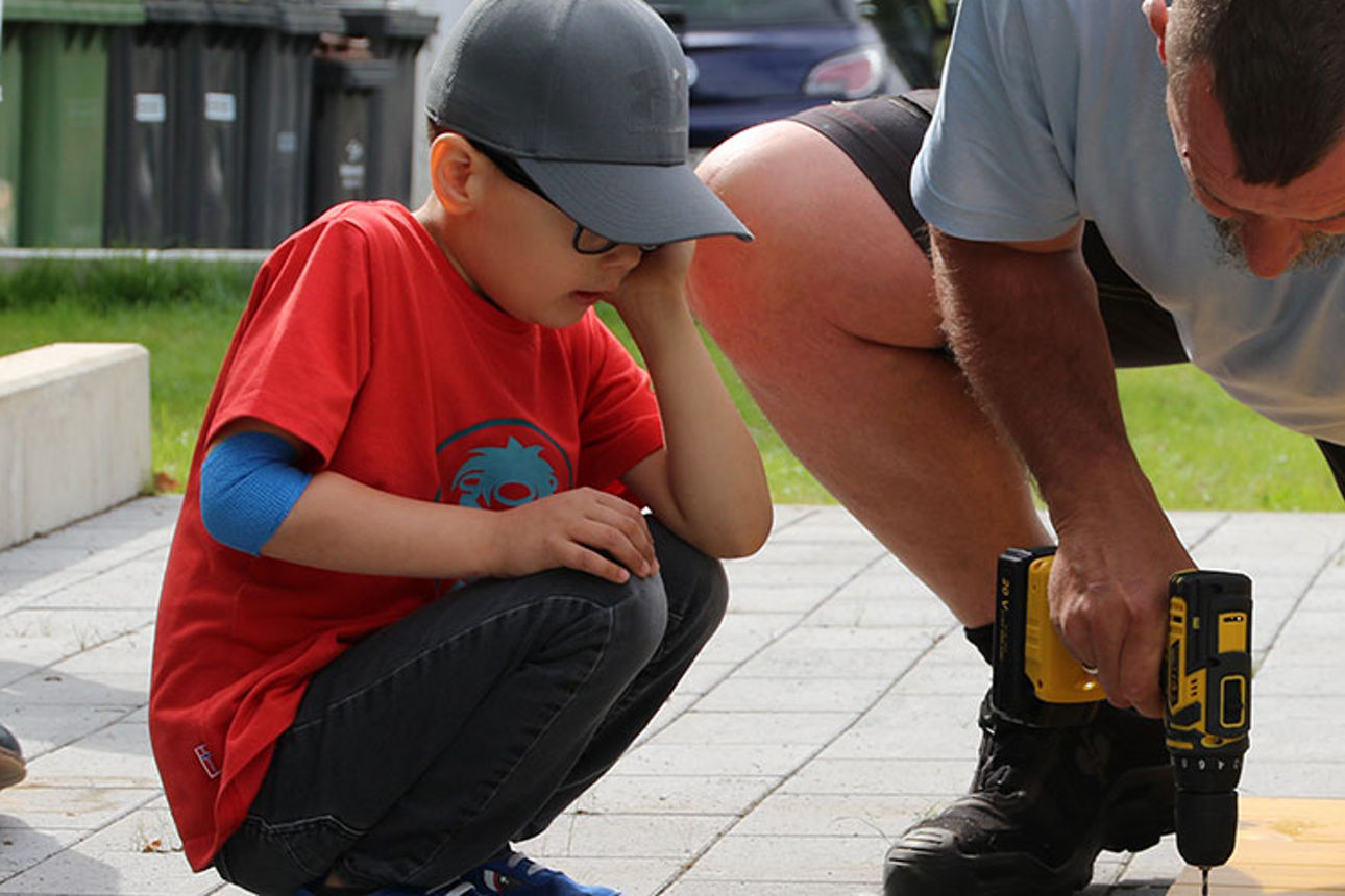 Ein Junge und ein Mann knien über einer Holzplatte. Der Mann hat einen Akkuschrauber in der Hand. Sie tragen Sommerkleidung.