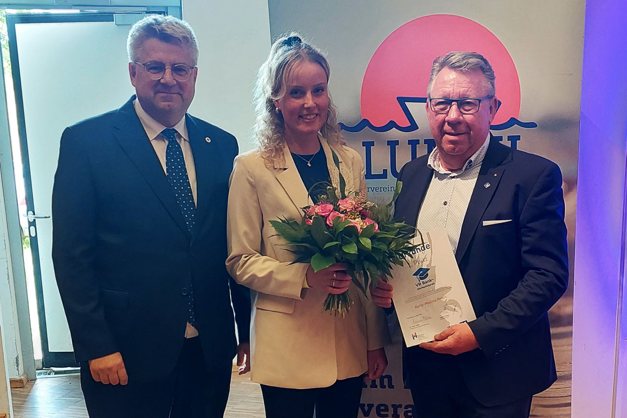 Eine junge Frau steht zwischen zwei Männern und wird mit einen Blumenstrauß und einer Urkunde geehrt.