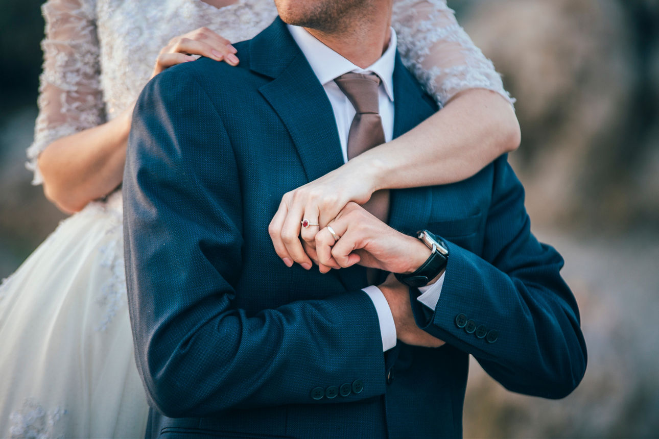 Braut im weißen Hochzeitskleid umarmt den Bräutigam mit ihrer Hand, an der sie einen Ring trägt.
