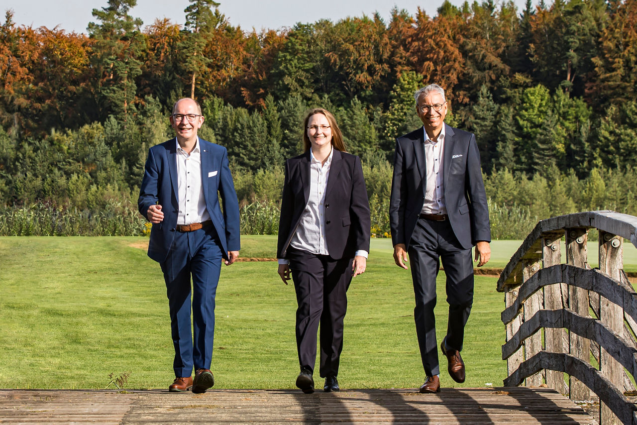 Die drei Vorstände, zwei Männer und eine Frau, laufen in einer Reihe über eine Brücke neben einer Wiese. Sie tragen Business Kleidung.