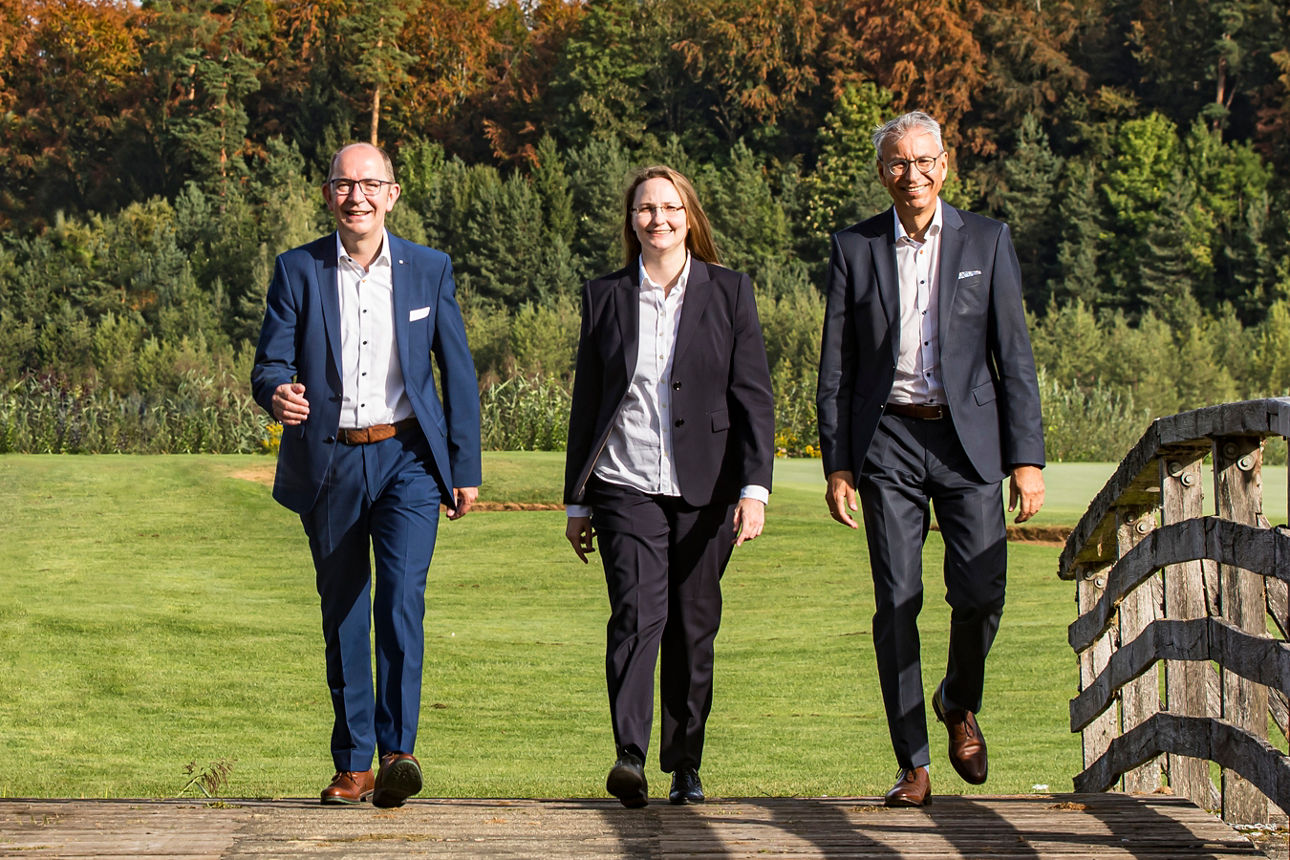 Die drei Vorstände, zwei Männer und eine Frau, laufen in einer Reihe über eine Brücke neben einer Wiese. Sie tragen Business Kleidung.