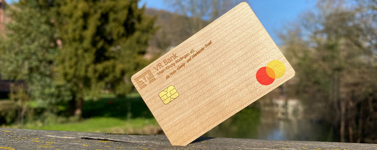 Eine Mastercard aus Holz mit dem Logo der VR Bank Main-Kinzig-Büdingen, im Hintergrund eine Parkanlage
