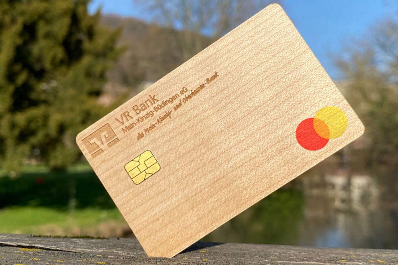 Eine Mastercard aus Holz mit dem Logo der VR Bank Main-Kinzig-Büdingen, im Hintergrund eine Parkanlage