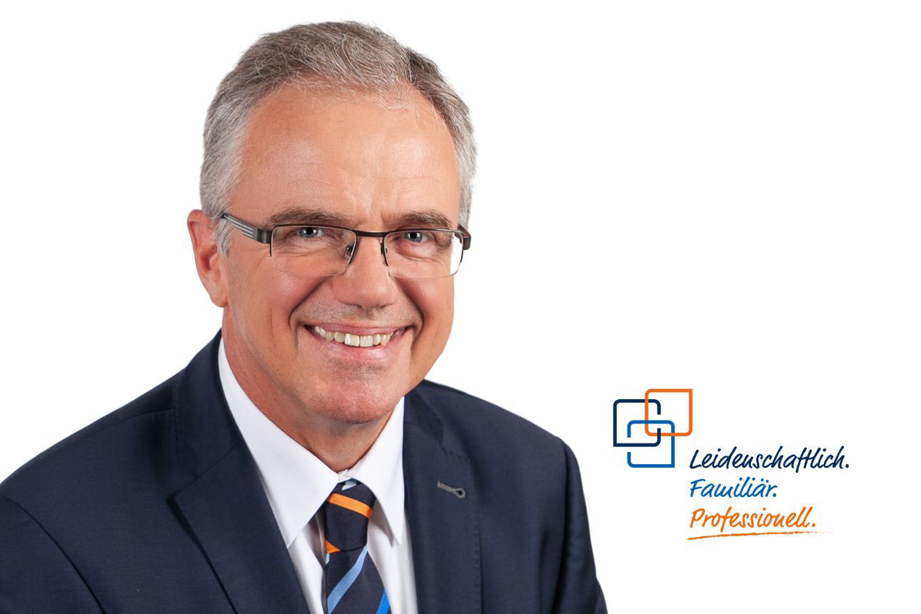 Horst Kürzeder, ein Mann mit grauem Haar und Brille, lächelt breit und trägt einen dunklen Anzug mit einer gestreiften blau-orangenen Krawatte.