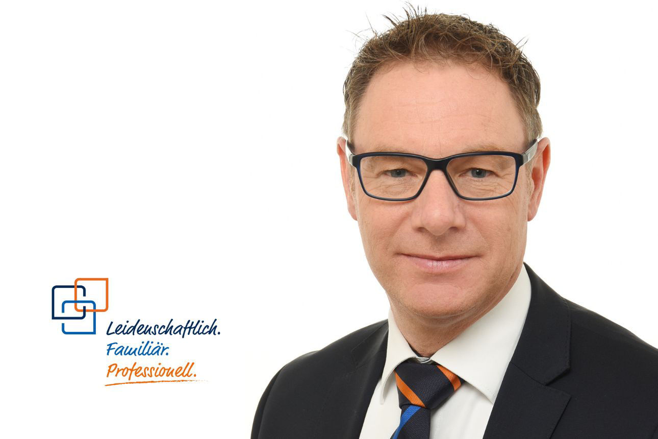Hubert Häusler, ein Mann mit Brille und kurzen, rötlich-braunen Haaren, trägt ein dunkles Sakko mit einem gestreiften Krawatten und blickt mit einem leichten Lächeln in die Kamera.