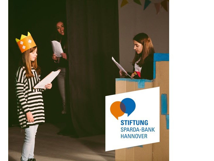 Zwei Mädchen sprechen mit Skripten in der Hand und mit Requisiten etwas vor; im Hintergrund schaut eine Frau lächelnd zu