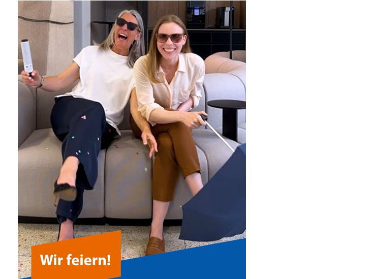 Zwei Frauen sitzen mit Sonnenbrillen auf einem Sofa und lachen herzhaft
